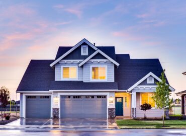 Modern two-storey suburban house with grey exterior, double garage, lit windows, and a landscaped lawn at dusk.